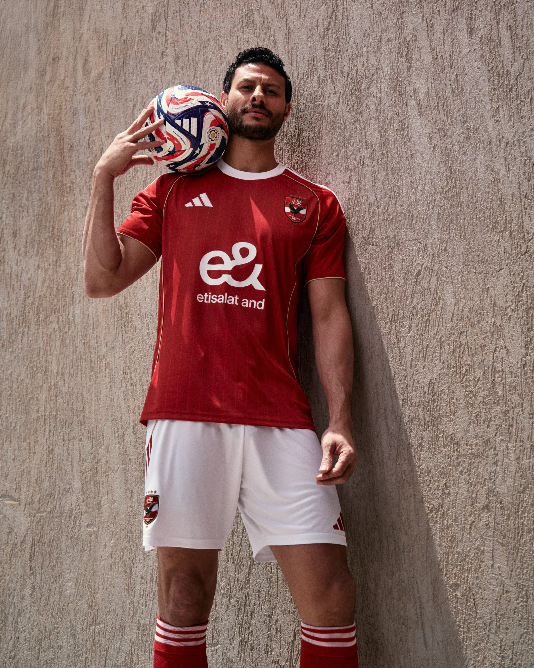 Footballer in a red Al Ahly kit holding a match ball on his shoulder against a stone wall