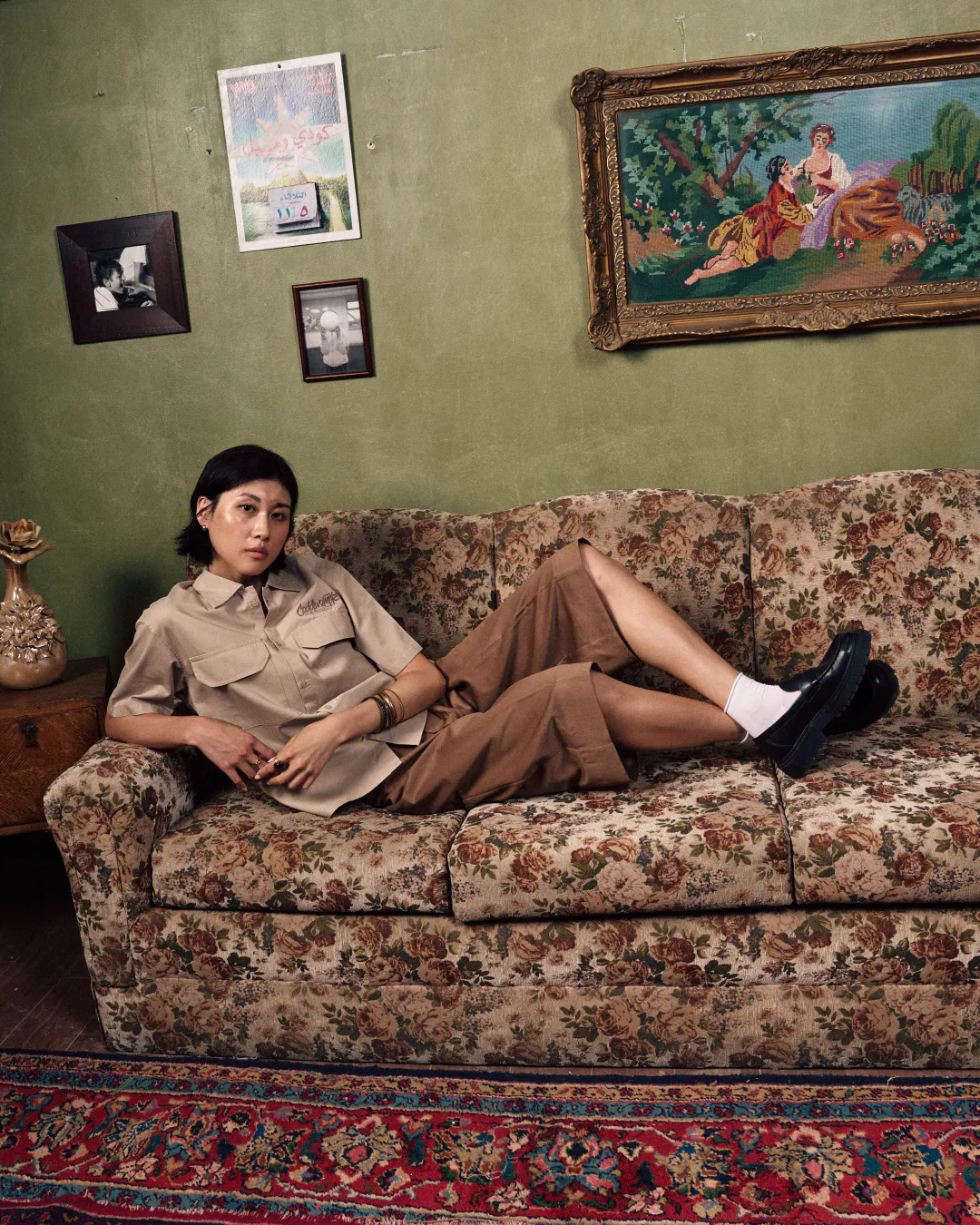 Model in a beige shirt and brown trousers reclining on a floral sofa in a green room