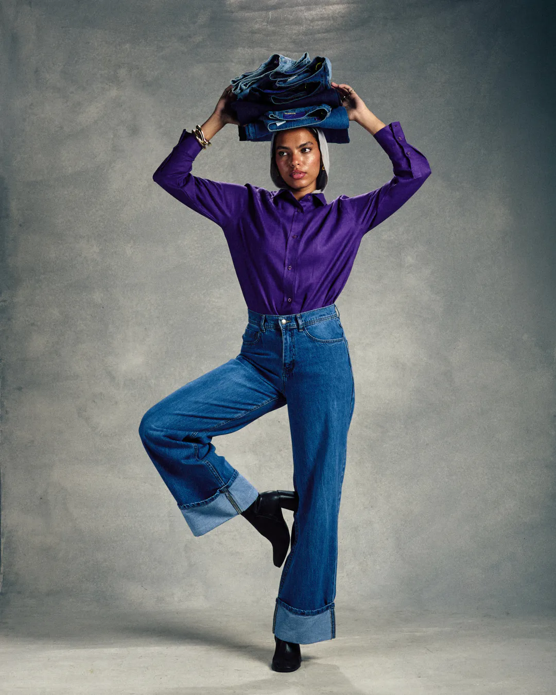 Woman in a purple shirt and wide-leg jeans balancing a stack of denim on her head